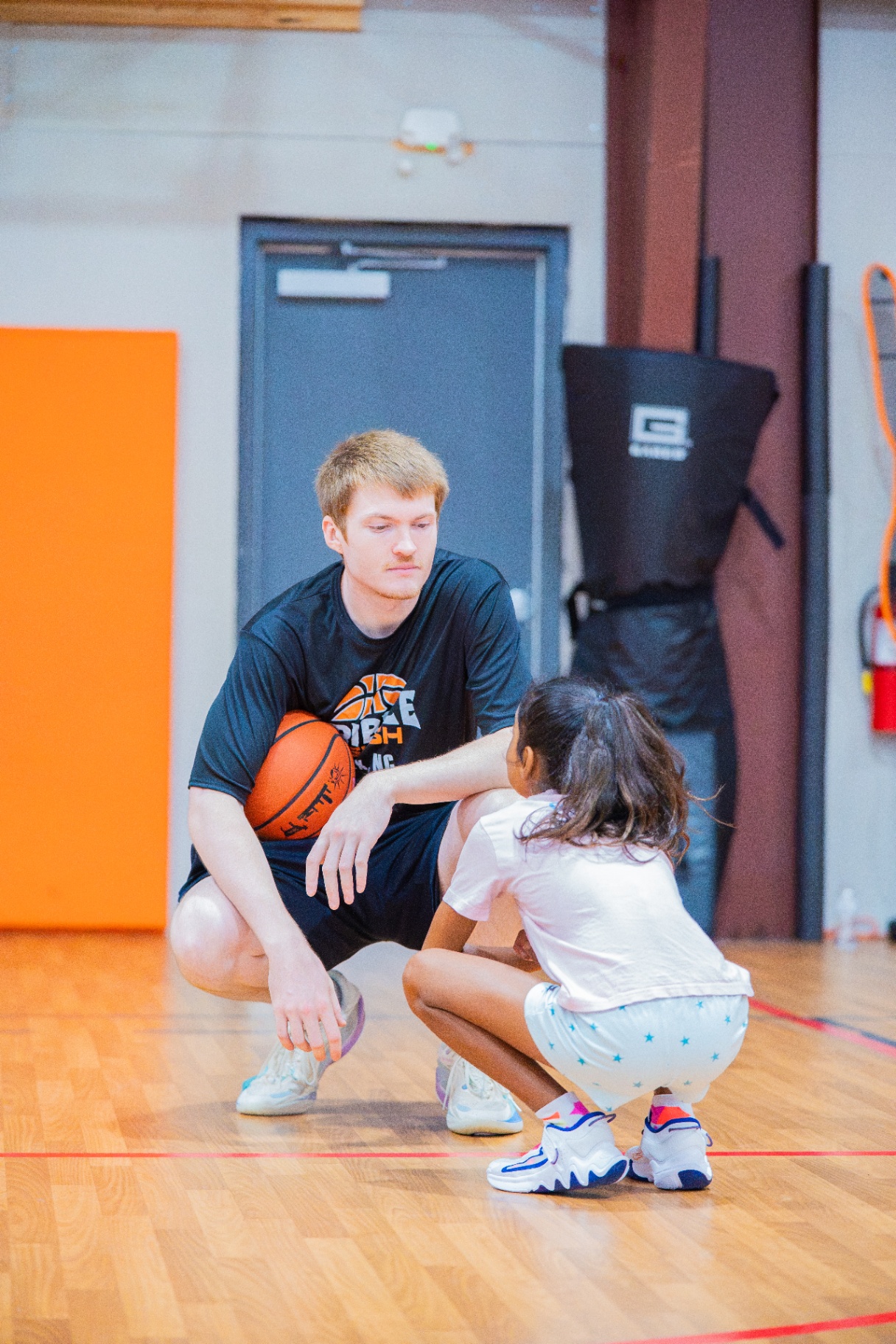 Coach crouched low with a basketball while speaking to a young player at court level