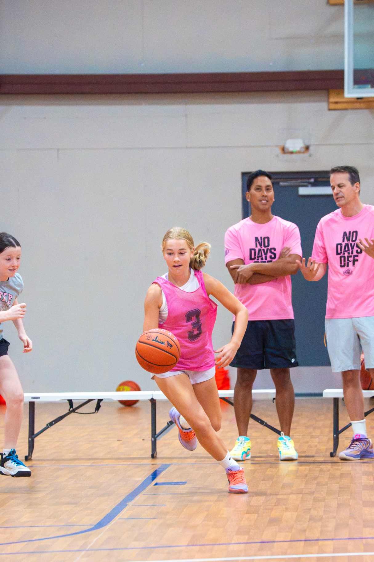 Player in a pink #3 pinnie pushing the ball in transition as coaches look on