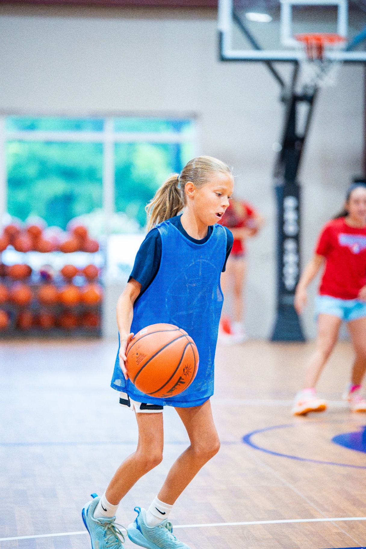 Young player in a blue pinnie controlling a dribble in front of a rack of basketballs